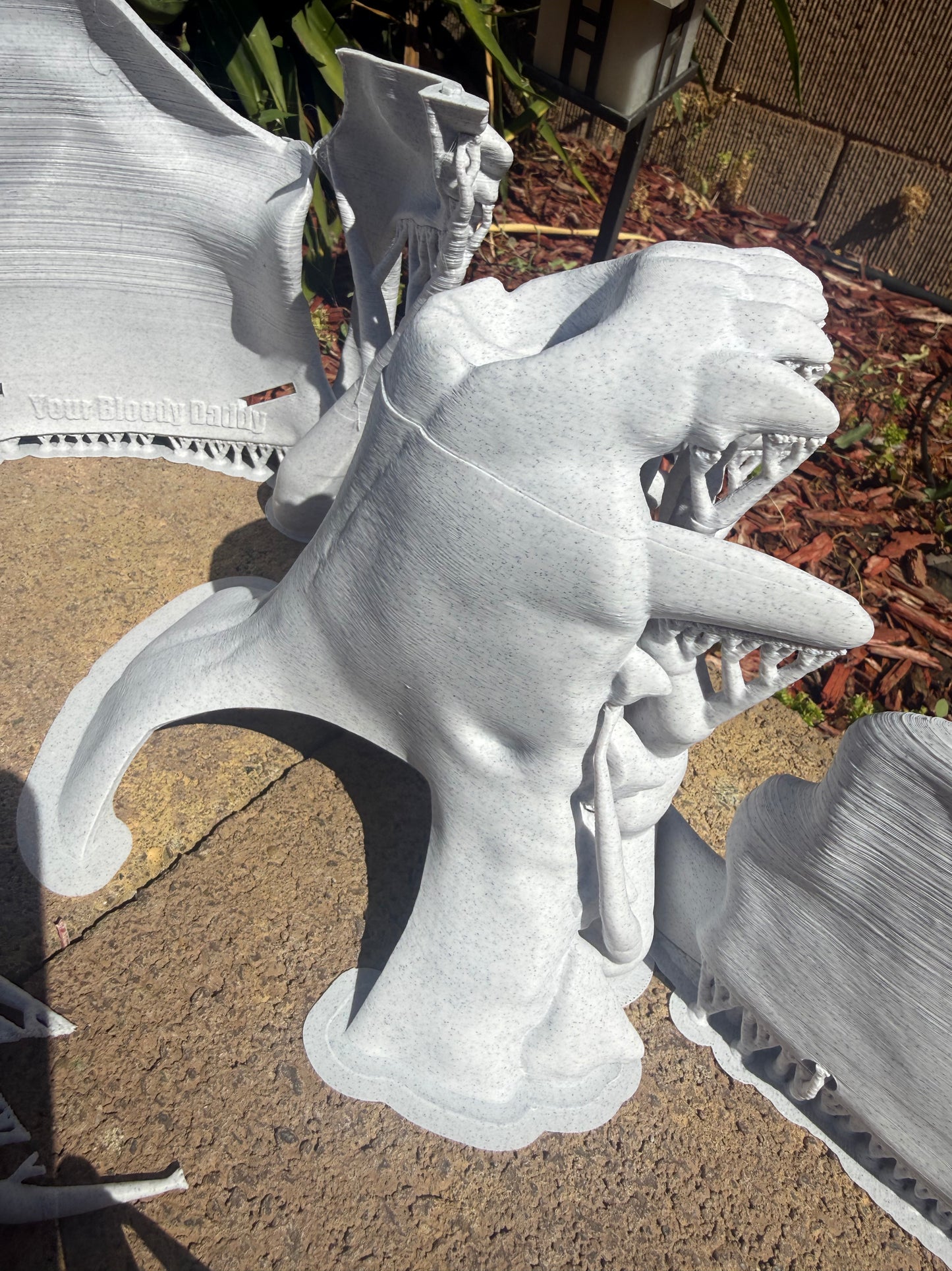 A light gray 3D-printed k9 skull sculpture displayed outdoors on a concrete patio with plants in the background.
