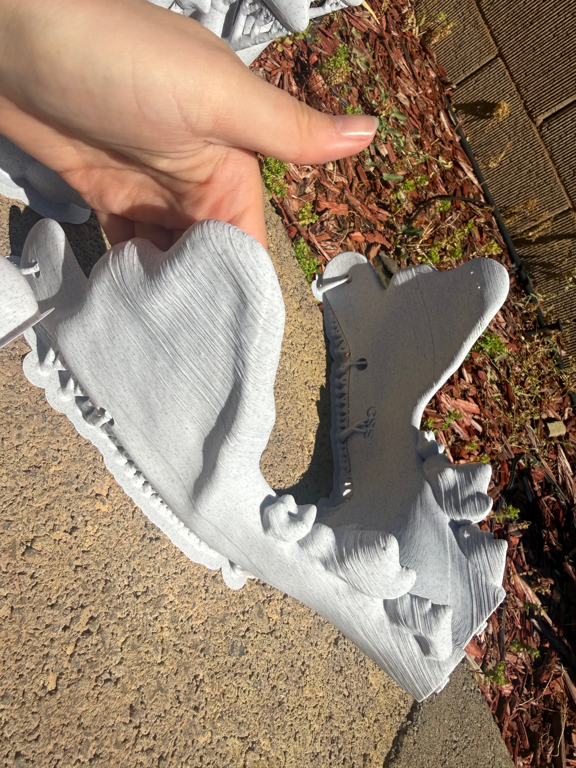 Half of the jaw of a light gray 3D-printed k9 skull sculpture displayed outdoors on a concrete patio with plants in the background.